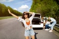 Young woman trying to wave down car, asking for help, her friends trying to fix broken vehicle, having flat tire on road Royalty Free Stock Photo