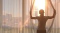 Young woman uncover the big window and looking out her apartment on the city buildings. Sunrise in the city. Royalty Free Stock Photo