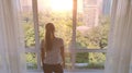 Young woman uncover the big window and looking out her apartment on the city buildings. Sunrise in the city. Royalty Free Stock Photo