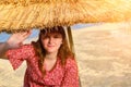 A young woman under a beach straw umbrella, copy space. Girl in a red dress on the evening in sunset near the blue sea. Female