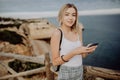 Young woman updating her social network status while relaxing at the rock beach with panorama view. Travel concept.