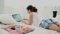 Young woman using laptop during breakfast lying on white bed at home. Brunette girl typing on pc computer. Royalty Free Stock Photo