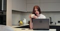 Young woman using laptop computer at kitchen. Female using a computer to browse on internet and drinking coffee at home Royalty Free Stock Photo