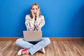 Young woman using laptop at home sitting on the floor afraid and shocked, surprise and amazed expression with hands on face Royalty Free Stock Photo