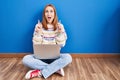 Young woman using laptop at home sitting on the floor amazed and surprised looking up and pointing with fingers and raised arms Royalty Free Stock Photo