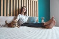 Young woman using laptop to work, lying on the bed with a pet cat in the bedroom, . Flexible hours and remote work.