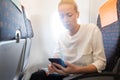 Young woman using mobile phone on airplane. Female traveler reading on her phone on the aircraft seat near the window Royalty Free Stock Photo