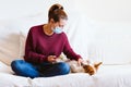 young woman using mobile phone, cute small dog besides. Sitting on the couch, wearing protective mask. Stay home concept during Royalty Free Stock Photo