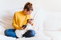 Young woman using mobile phone, cute small dog besides. Sitting on the couch, wearing protective mask. Stay home concept during Royalty Free Stock Photo