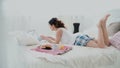 Young woman using wireless technology lying on a white bed at home. Girl chatting on laptop during breakfast. Royalty Free Stock Photo