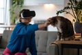 Young woman in VR glasses touches domestic cat and strokes animal head in real life in apartment