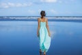 Young woman walking barefoot on empty beach. Full body portrait. Slim Caucasian woman wearing long dress. View from back. Summer