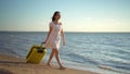 Young woman walks along the beach of the sea with a yellow suitcase. A girl in a white dress walks barefoot on the sand Royalty Free Stock Photo