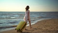 Young woman walks along the beach of the sea with a yellow suitcase. A girl in a white dress walks barefoot on the sand. Royalty Free Stock Photo