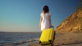 Young woman walks along the beach of the sea with a yellow suitcase. A girl in a white dress walks barefoot on the sand. Royalty Free Stock Photo