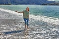 Young woman walks barefoot on beach in surf zone.