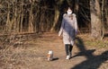 Young woman in warm jacket, wearing virus face mouth mask, walks her dog on country forest road. Masks are mandatory outside home