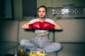 Young woman watch boxing on tv at night. Sitting with legs crossed and hands in boxing gloves. Serious concentrated