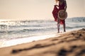 Young woman in waving red summer dress walking on beach holding fashionable straw hat. Vacation, holiday, lifestyle concept Royalty Free Stock Photo