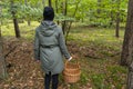 A young woman wearing a black cap and olive coat stands backwards in the woods holding a wicker basket to collect mushrooms.