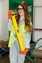 Young woman wearing denim jumpsuit and glasses with long hair holding in hands spirit level and smiling. Royalty Free Stock Photo