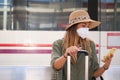 Young woman wearing face mask and explorer hat, using her smartphone while is waiting for the train at a train station Royalty Free Stock Photo