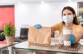 Young woman wearing face mask while serving takeaway breakfast and coffee inside cafeteria restaurant - Worker preparing delivery