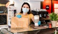 Young woman wearing face mask while serving takeaway breakfast and coffee inside cafeteria restaurant - Worker preparing healthy