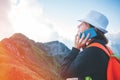 Young woman wearing a hat and backpack talking on the phone, sitting high in mountains at sunset