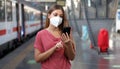 Young woman wearing KN95 FFP2 face mask waiting train at the station. Casual woman using smart phone in train station