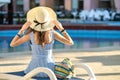 Young woman wearing light summer dress and yellow straw hat sitting outside near hotel swimming pool on summer sunny day Royalty Free Stock Photo