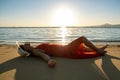 Young woman wearing long red dress and straw hat laying on sand beach at sea shore enjoying view of rising sun in early summer Royalty Free Stock Photo