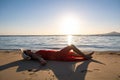 Young woman wearing long red dress and straw hat laying on sand beach at sea shore enjoying view of rising sun in early summer Royalty Free Stock Photo