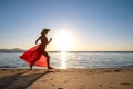 Young woman wearing long red dress and straw hat running on sand beach at sea shore enjoying view of rising sun in early summer Royalty Free Stock Photo