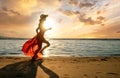 Young woman wearing long red dress and straw hat running on sand beach at sea shore enjoying view of rising sun in early summer Royalty Free Stock Photo