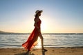 Young woman wearing long red dress and straw hat standing on sand beach at sea shore enjoying view of rising sun in early summer Royalty Free Stock Photo