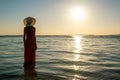 Young woman wearing long red dress and straw hat standing in sea water at the beach enjoying view of rising sun in early summer Royalty Free Stock Photo