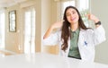 Young woman wearing medical coat at the clinic as therapist or doctor smiling confident showing and pointing with fingers teeth