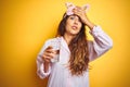 Young woman wearing pajama and sleep mask drinking water over yellow isolated background stressed with hand on head, shocked with Royalty Free Stock Photo