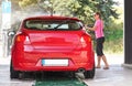 Young woman wearing pink t shirt and sunglasses washing her red car in self serve carwash, view from back