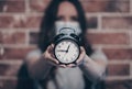 A young woman wearing a protective medical mask against coronavirus on her face, holding a alarm clock, on brick blurred