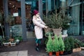 Young woman wearing protective medical mask in faux fur coat picks small Christmas tree in wicker pot in city flower shop