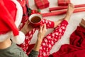 Young woman wearing santa hat and xmas pajamas sitting on the floor amongst wrapped christmas presents, drinking hot punch.