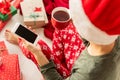 Young woman wearing santa hat and xmas pajamas sitting on the floor, drinking hot punch and shopping online on her phone. Royalty Free Stock Photo