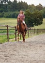 Young woman wearing shirt riding brown horse in sand paddock by wooden fence, hair moving in air because of speed Royalty Free Stock Photo