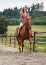 Young woman wearing shirt riding brown horse in sand paddock by wooden fence, hair moving in air because of speed Royalty Free Stock Photo