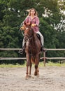 Young woman wearing shirt riding brown horse in sand paddock by wooden fence, hair moving in air because of speed Royalty Free Stock Photo