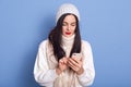 Young woman wearing sweater, scarf and cap, typing message, posing isolated over blue background, brunette lady wears red lipstick Royalty Free Stock Photo