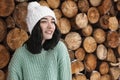 Young woman wearing warm sweater and hat near stack of firewood. Winter season