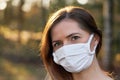 Young woman wearing white cotton mouth nose mask, blurred sunset lit trees in background, closeup face portrait